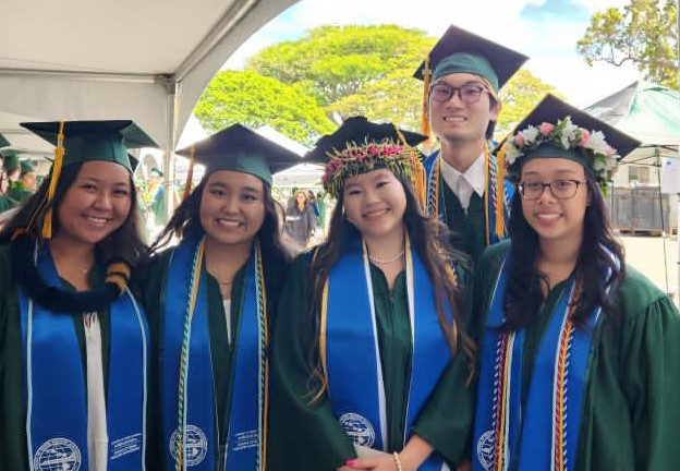 Image of five Hawaiʻi Tourism Hoʻoilina scholars (in caps and gowns) at their graduation from the University of Hawaiʻi. 