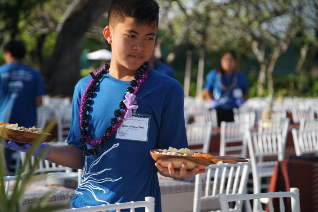 Image of a student participant with ClimbHI. He is carrying a plate of food in each hand and participating in hospitality training and learning to setup for events. 