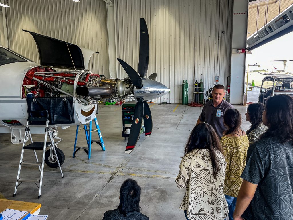 HTA Work Wise program. Image is in an airplane hangar with a propeller plane with the engine exposed. An instructor is speaking to students that are standing on the side. 