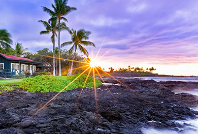 Image of a green-colored home with a red roof situated on the ocean with the sun setting in the background. 