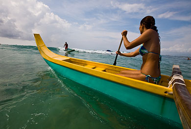 A canoe paddler in the ocean passes near a surfer riding a wave. 